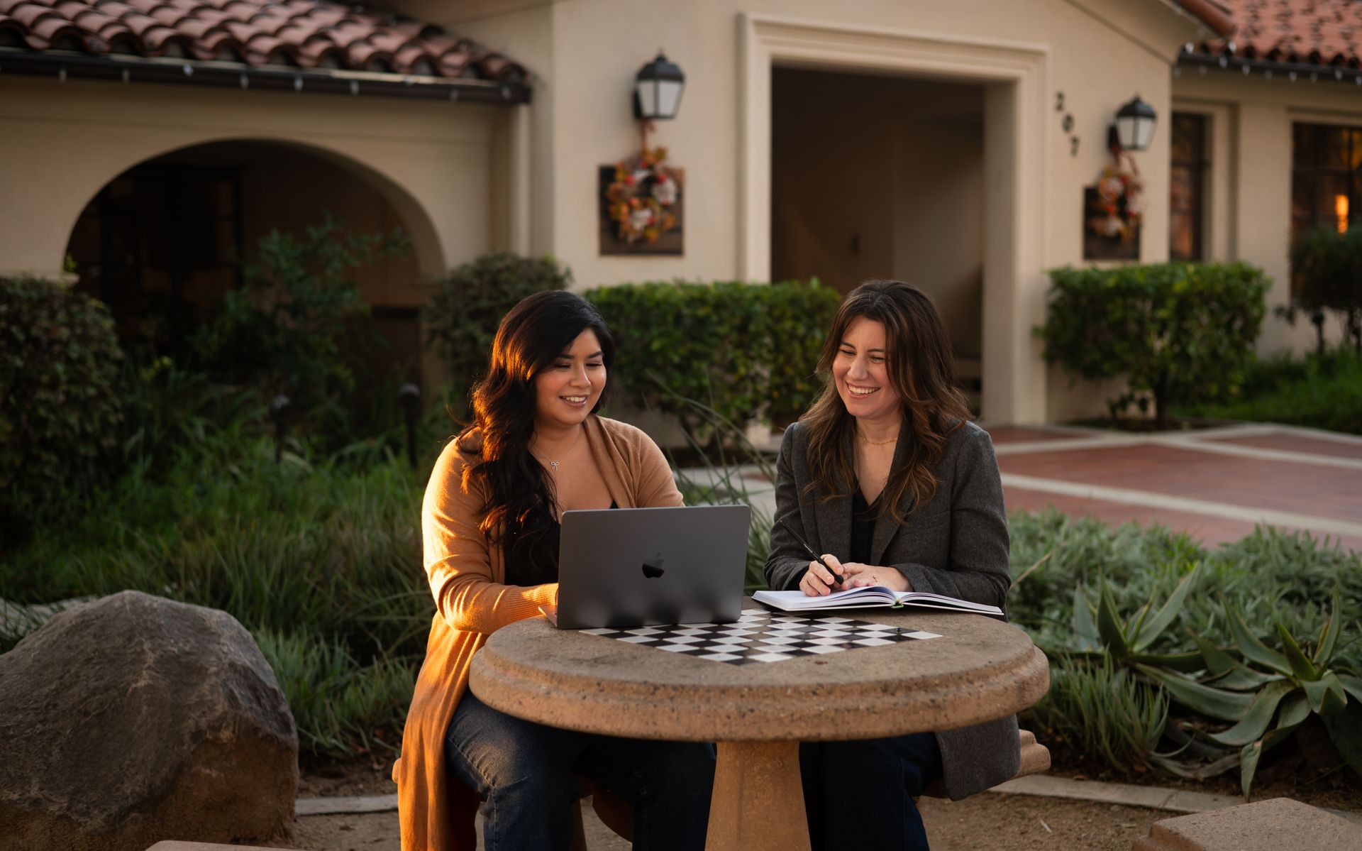 Two women sit at a stone table with a laptop, smiling. Outdoors, near a building.