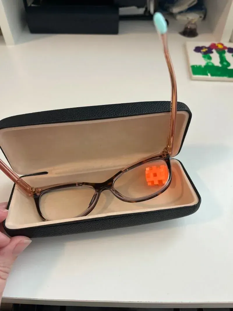 Eyeglasses in a black case on a white surface, glasses have brown frames, a blue end on one arm, and an orange square