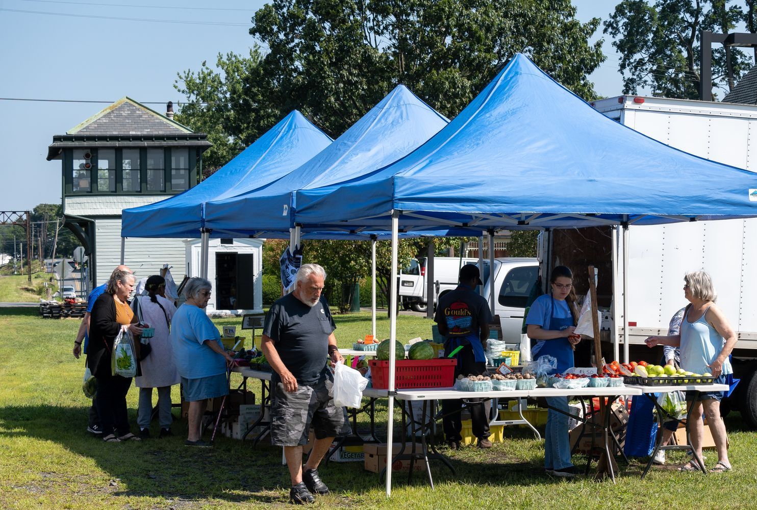 A group of people are standing under blue tents at a farmers market.