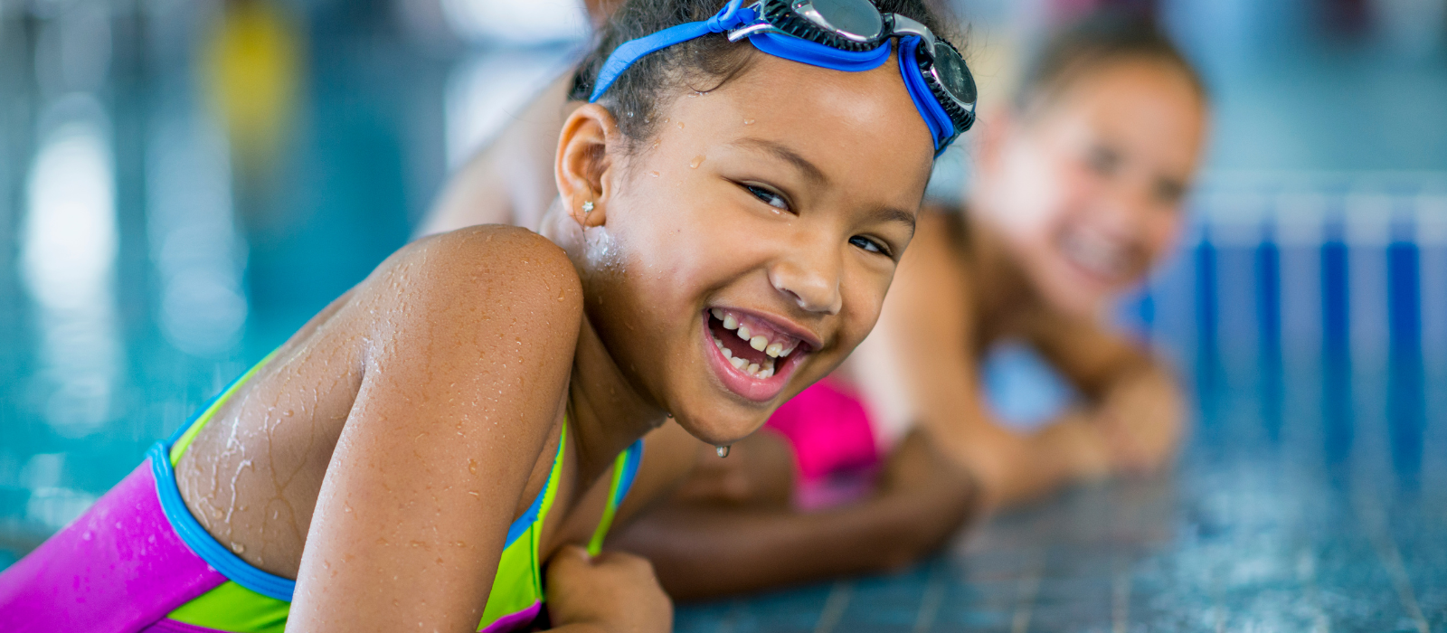 A little girl wearing goggles is laying on the edge of a swimming pool.