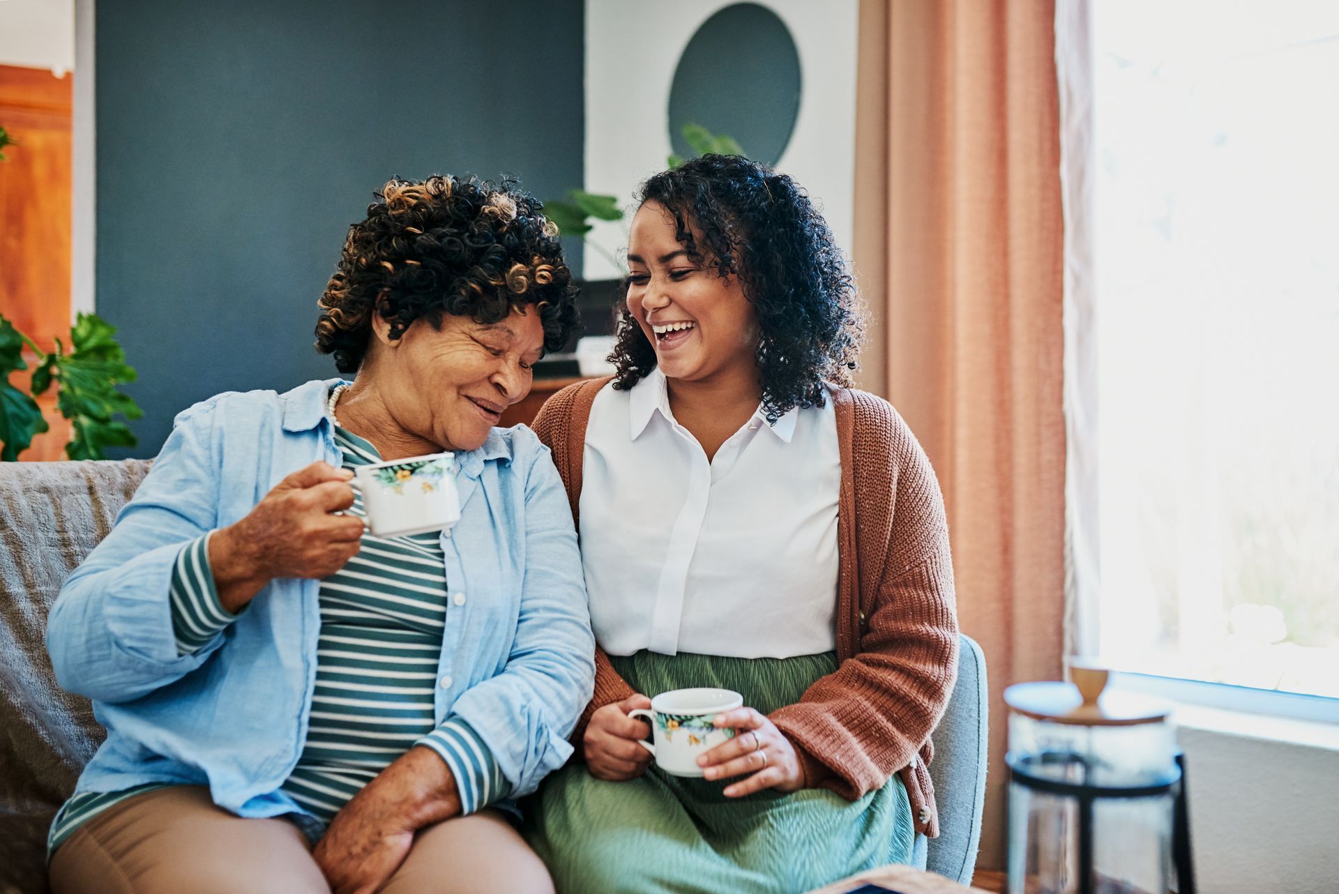 Two women are sitting on a couch drinking tea and laughing.