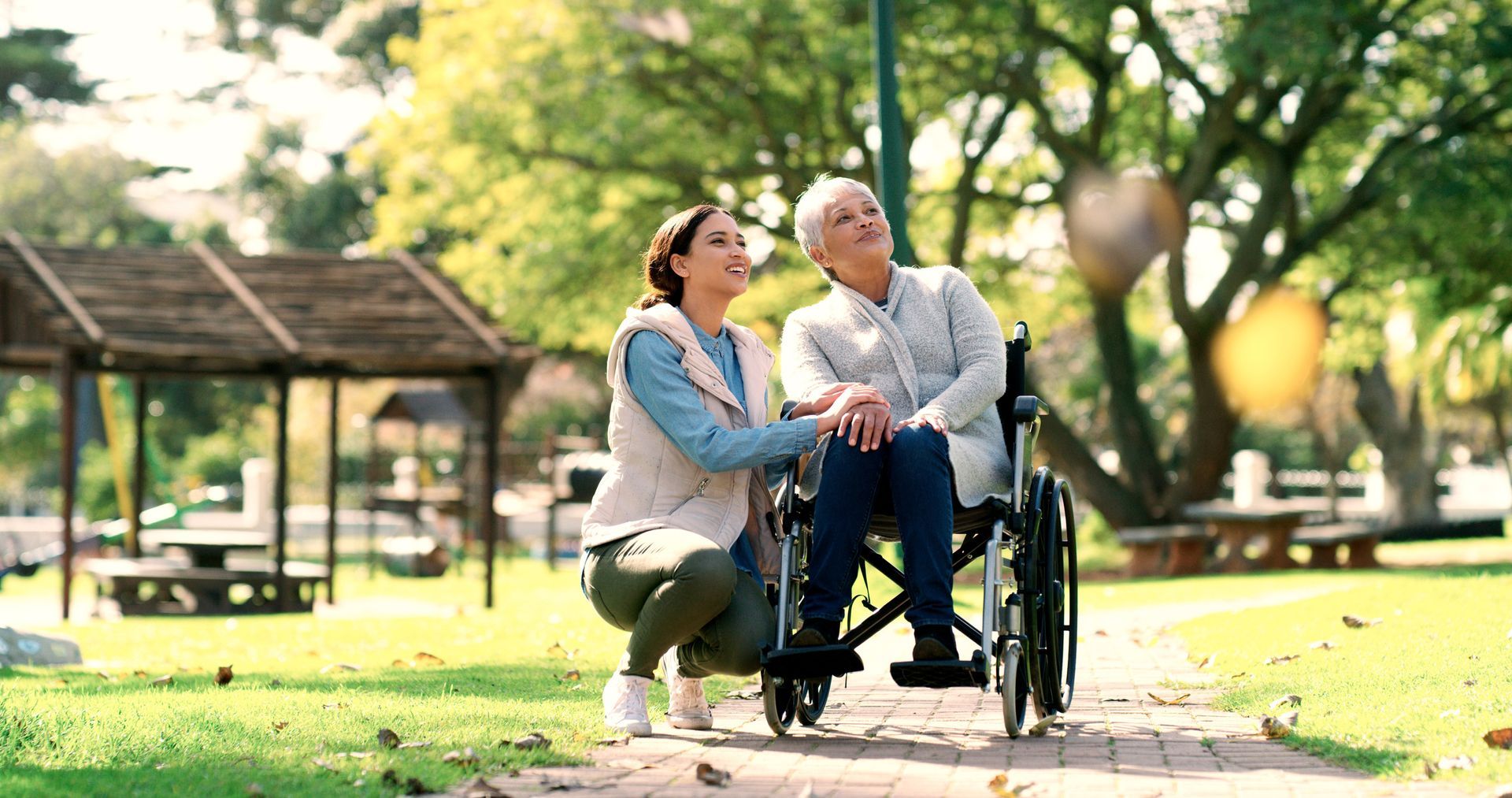 A woman is kneeling next to an elderly woman in a wheelchair in a park.
