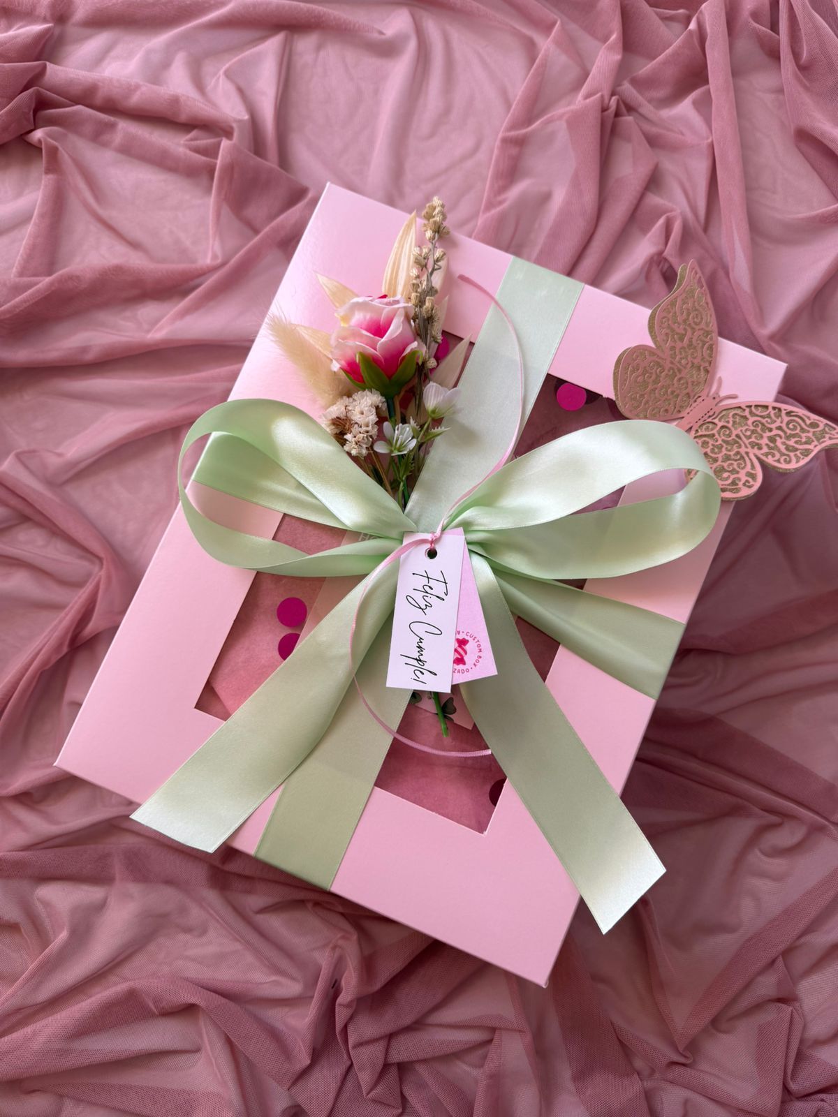 Una caja rosa con una cinta verde y flores dentro.