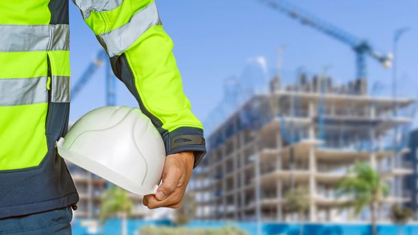 Operaio edile con in mano un casco bianco davanti a un edificio in costruzione.