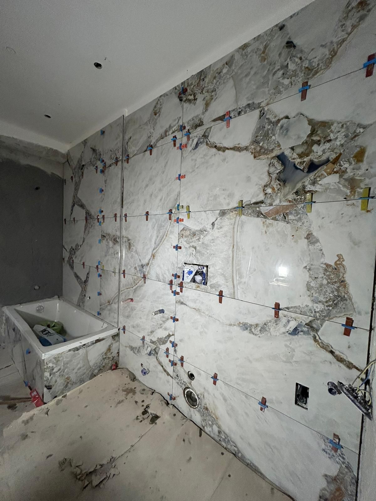 Bagno con pareti piastrellate con motivi in marmo in fase di installazione; vasca e prese elettriche visibili.