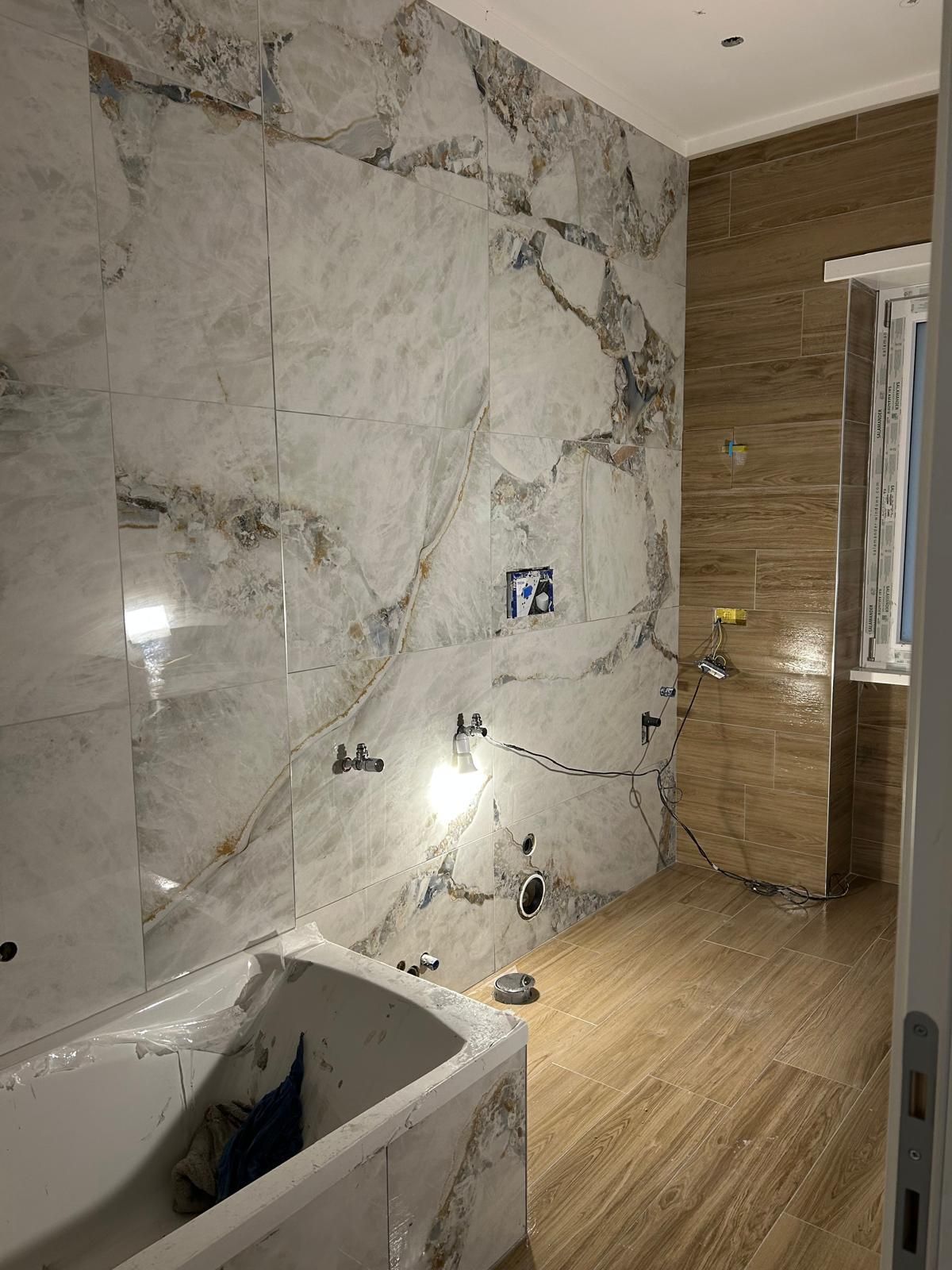 Bagno con pareti piastrellate con motivo marmo, pavimento in piastrelle effetto legno, vasca da bagno e sanitari grezzi.