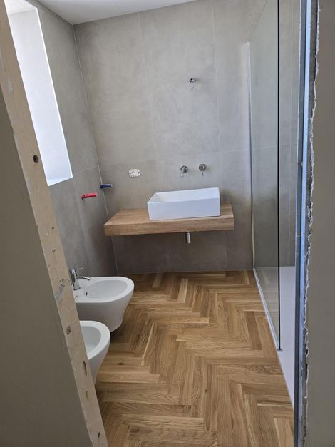 Bagno con pavimento in legno, lavandino sospeso in legno e doccia in vetro.