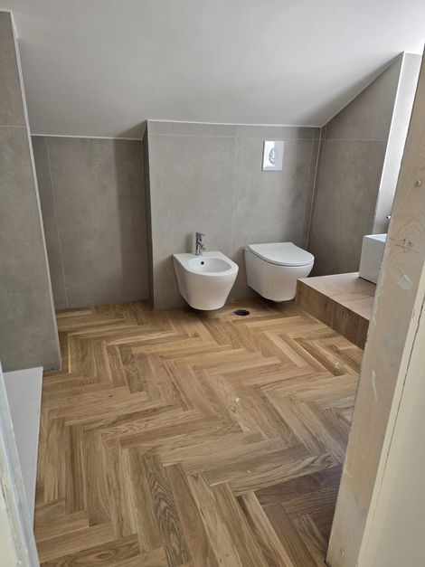 Bagno con pavimento in legno a spina di pesce, pareti grigie e WC e bidet bianchi.