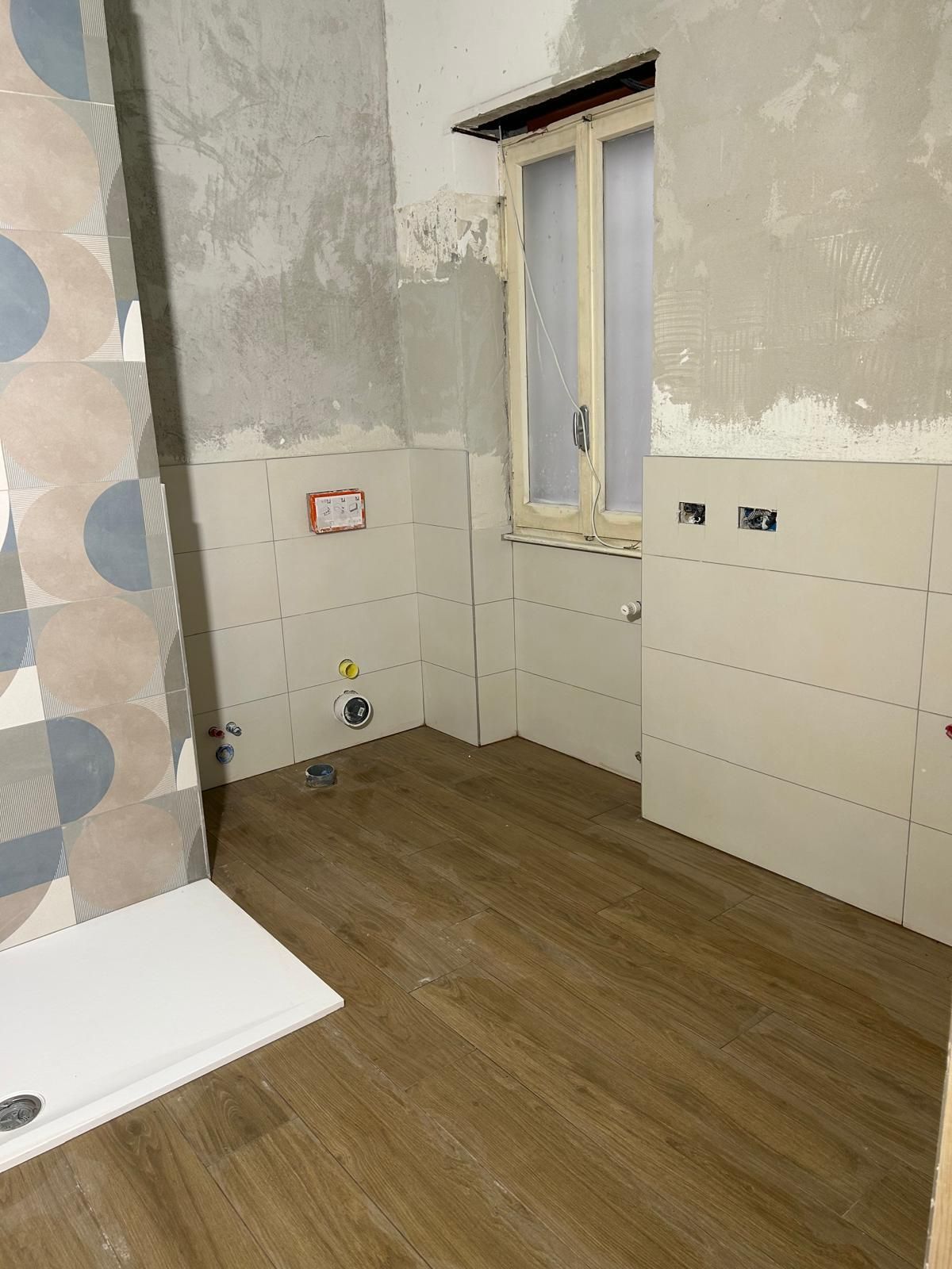 Ristrutturazione del bagno: parete piastrellata bianca e blu, finestra, pavimento in legno e piatto doccia.