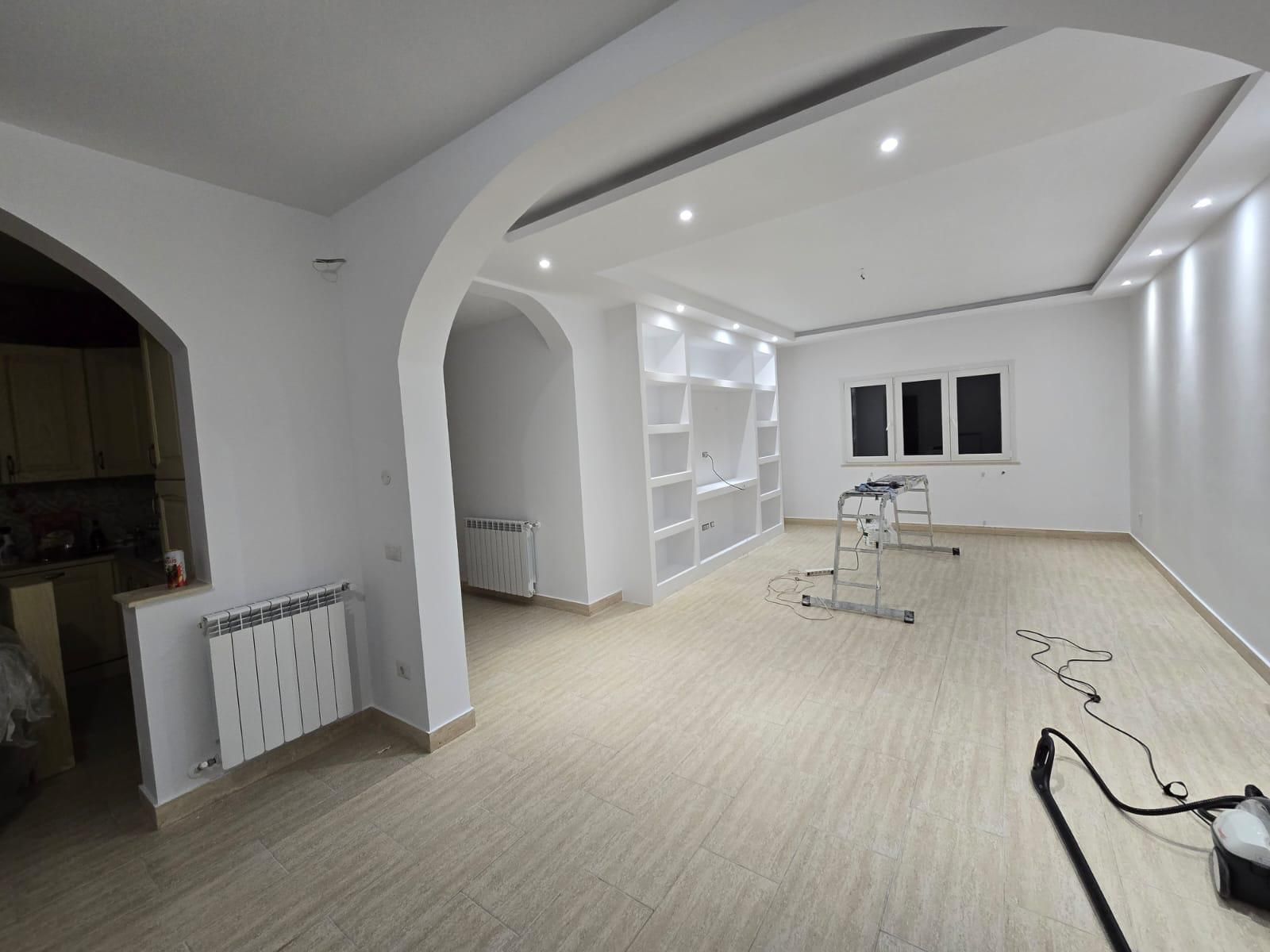 Stanza vuota con pareti bianche, archi, scaffali incorporati e pavimento effetto legno; sono presenti attrezzi da costruzione.