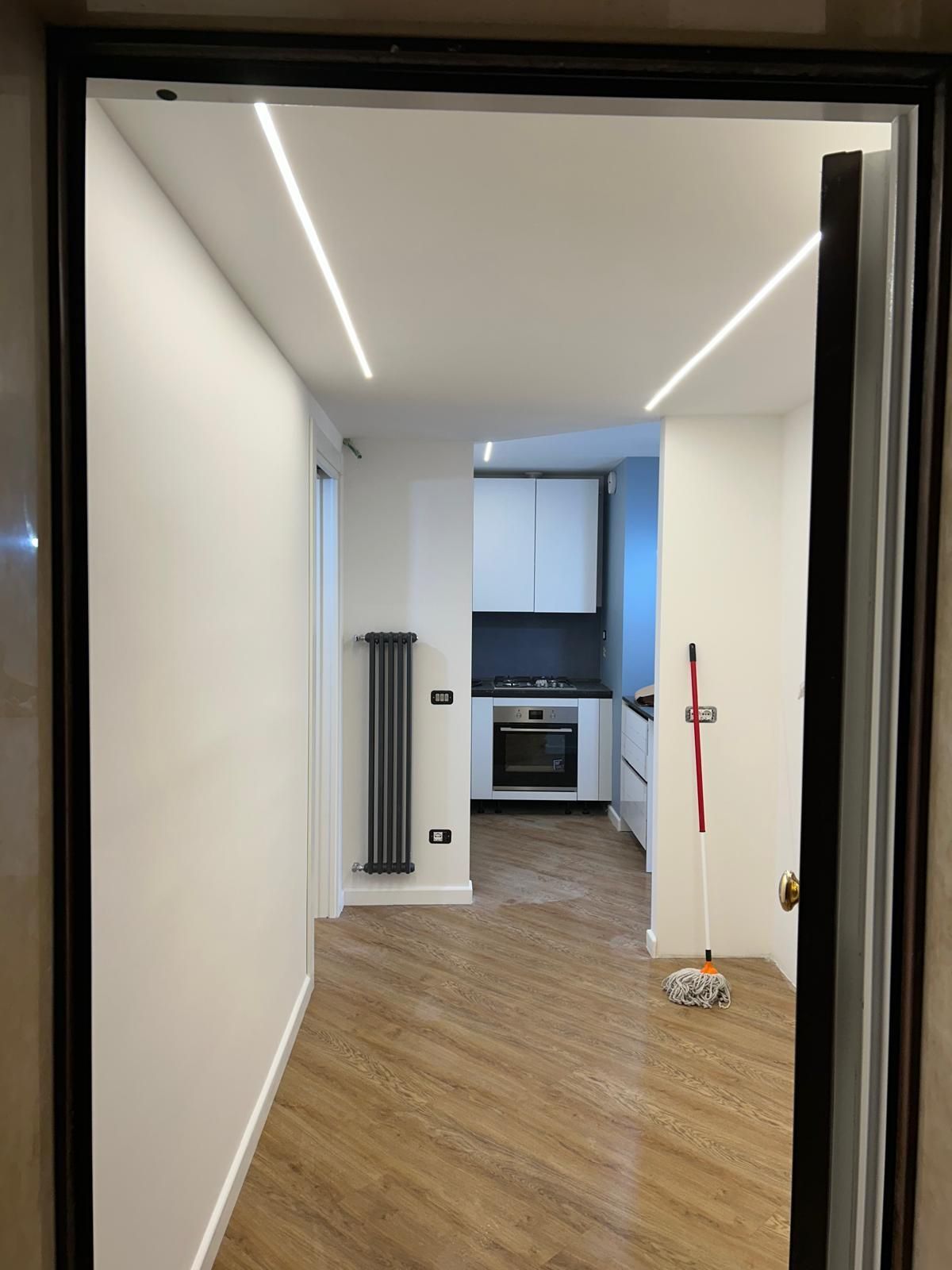 Corridoio che conduce alla cucina con mobili bianchi, termosifone grigio e pavimento in legno.