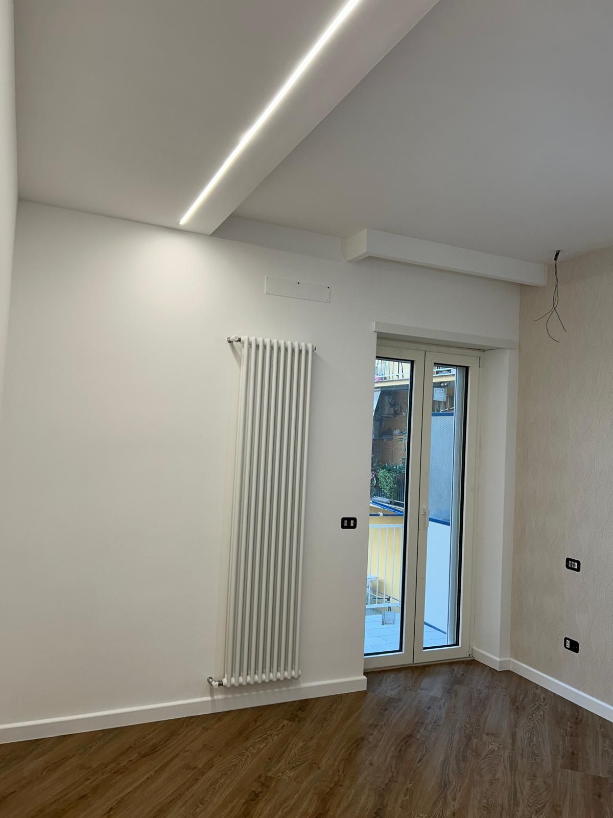 Stanza con pareti bianche, termosifone, porta che dà sul giardino e illuminazione da incasso; pavimento in legno.