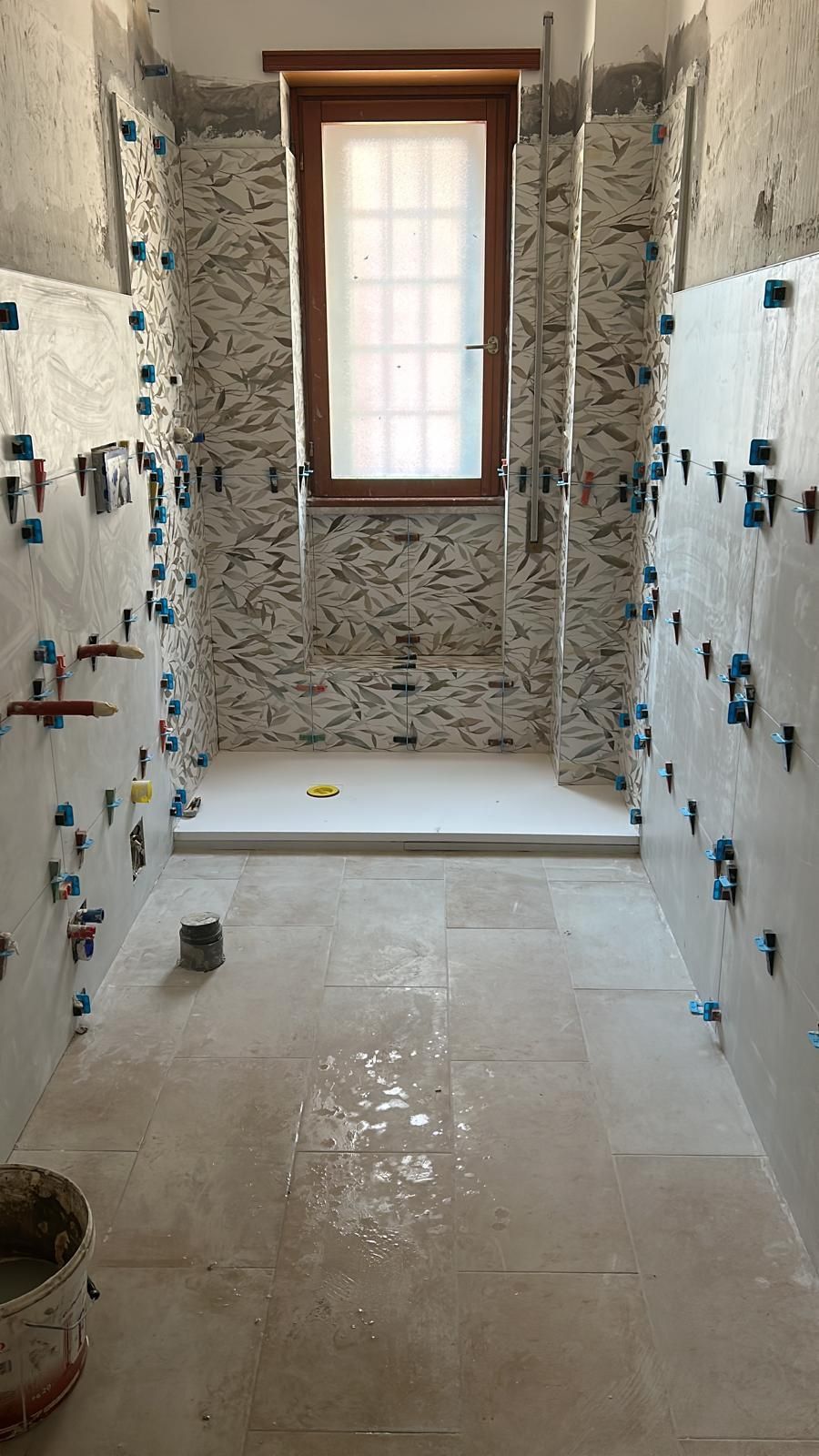 Ristrutturazione del bagno in corso. Installazione delle piastrelle in corso; doccia, finestra e pareti grezze visibili.