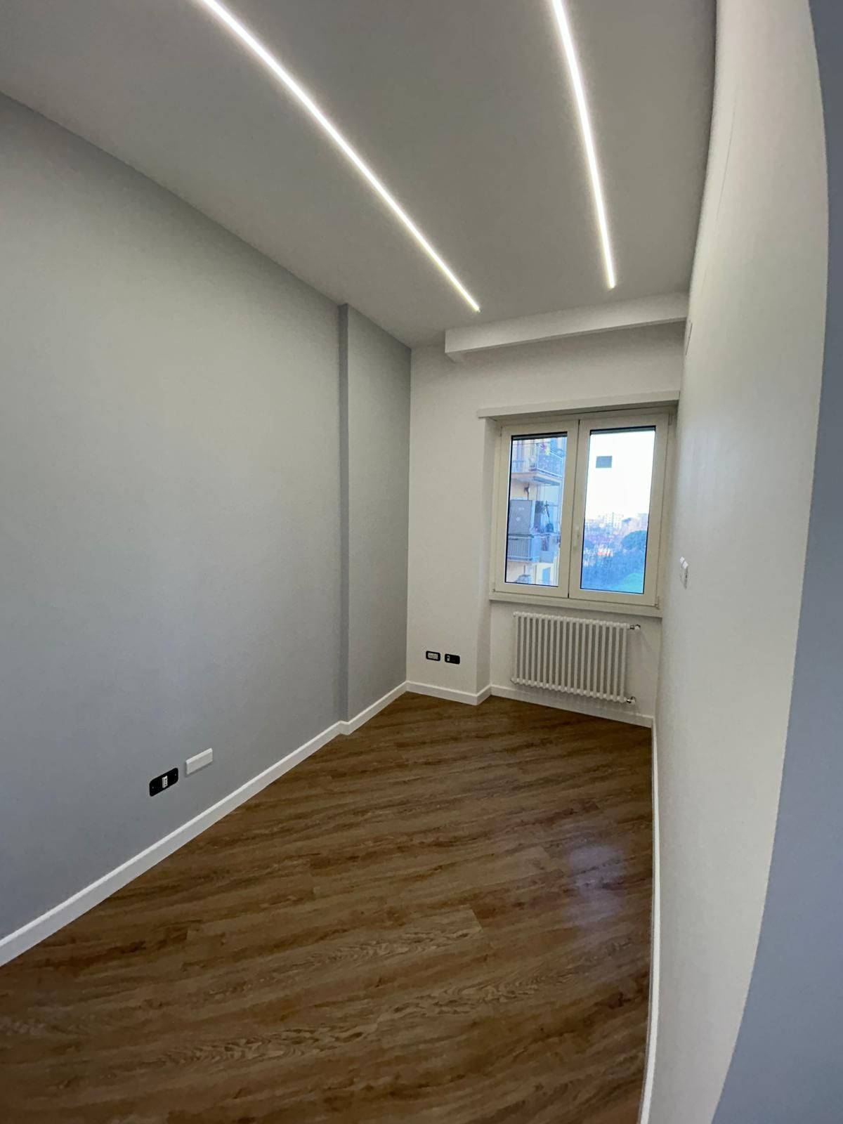 Stanza vuota con pavimento effetto legno, pareti grigie, finestra e luci al soffitto.