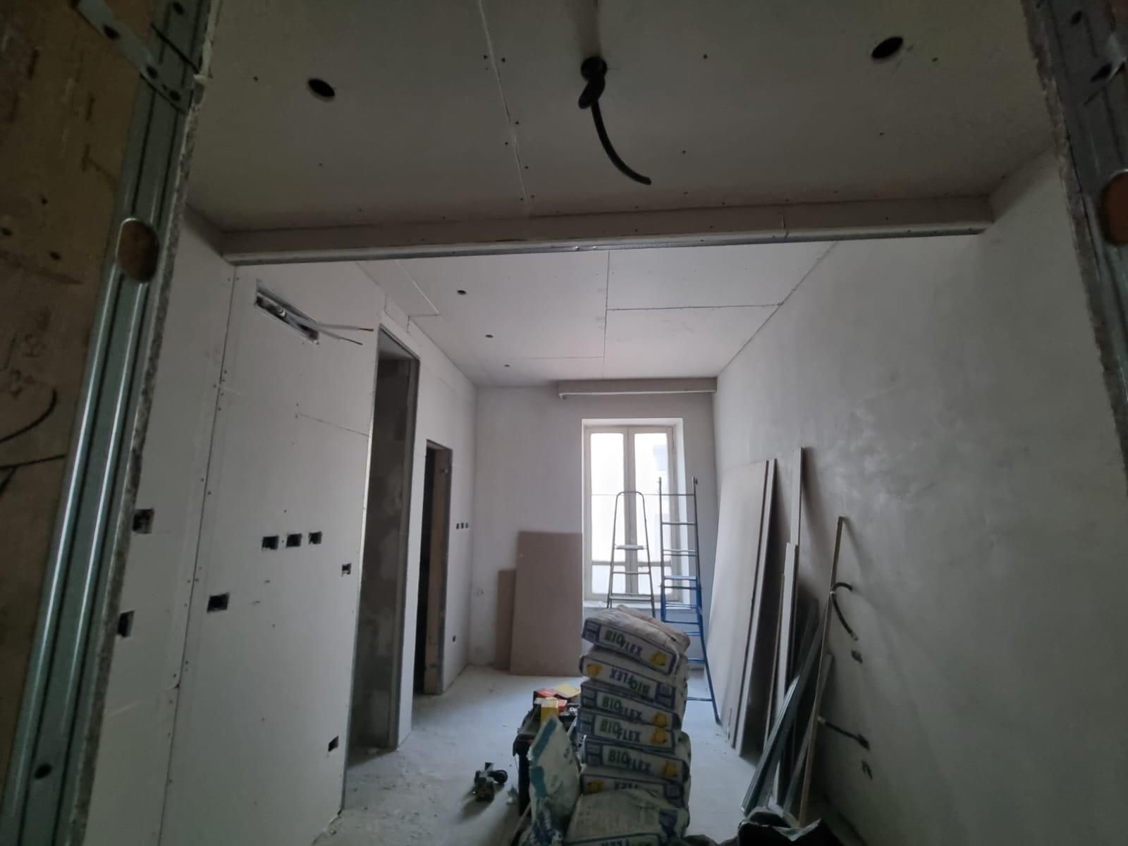 Interno in costruzione con cartongesso e sacchi di cemento; una finestra si trova in fondo al corridoio.