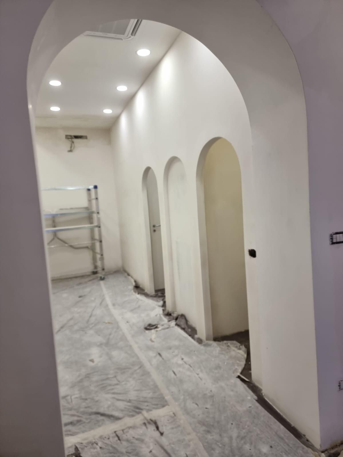Vista interna di un corridoio con porte ad arco e illuminazione da incasso; pavimento grezzo, pareti bianche.