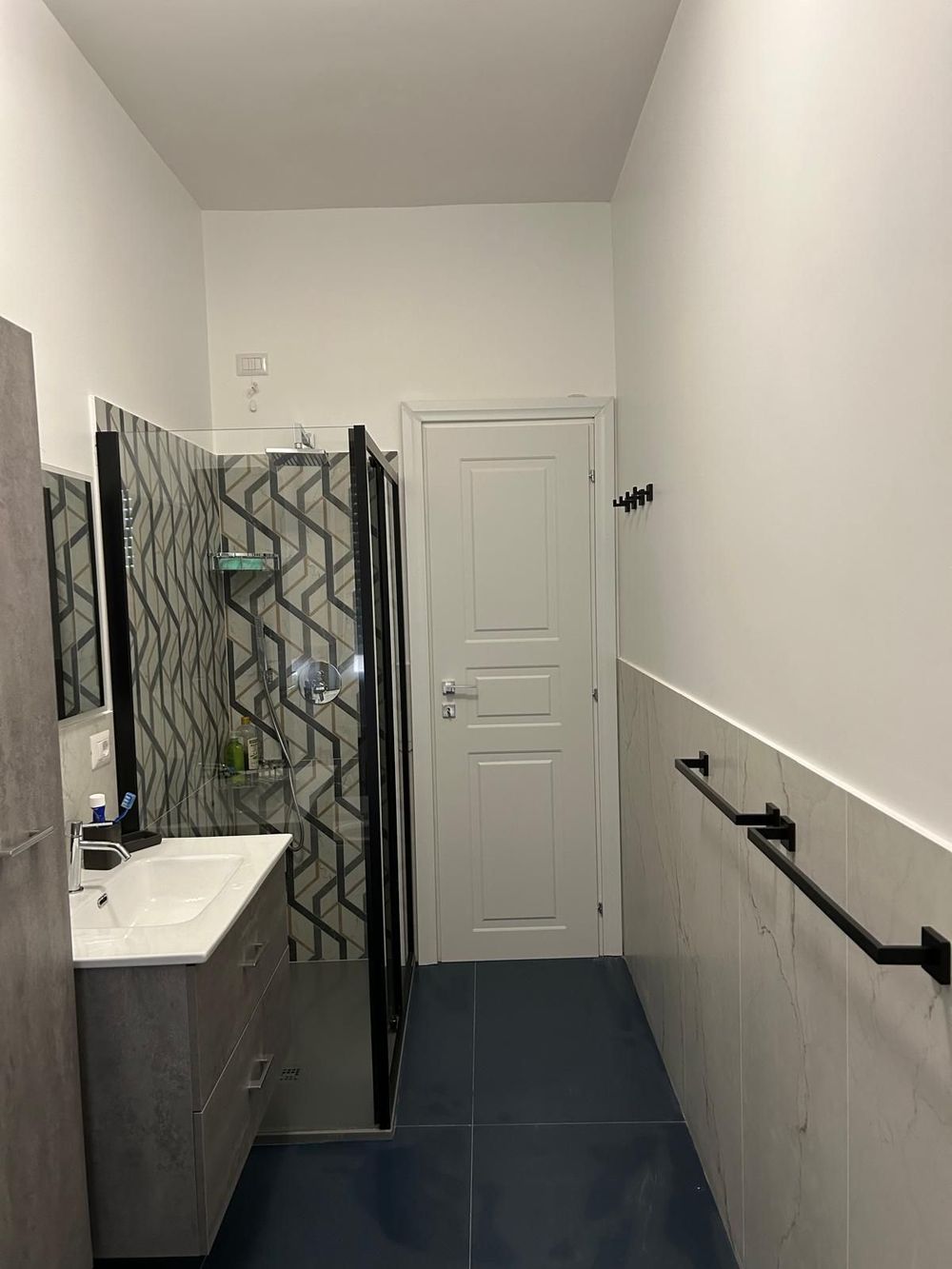 Bagno stretto con mobiletto grigio, doccia, porta bianca e portasciugamani neri.
