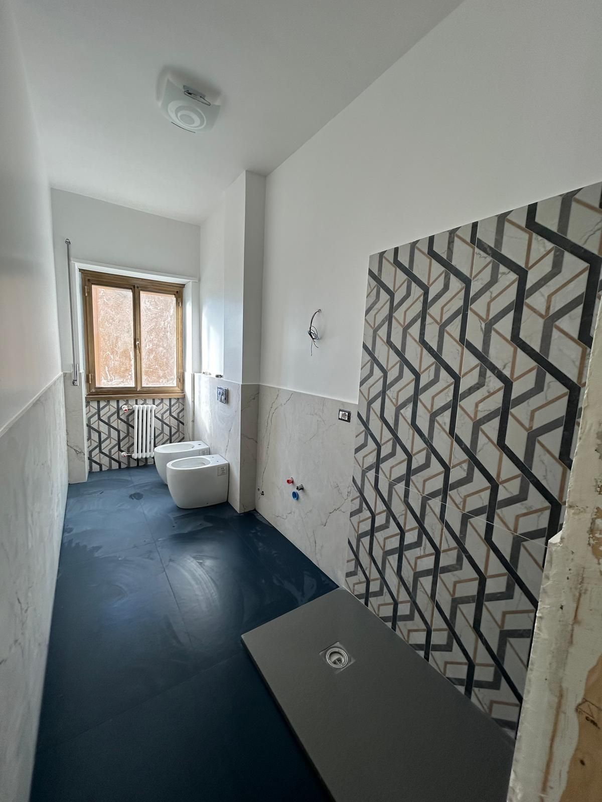 Bagno in costruzione: piastrelle con motivi geometrici, pavimento blu scuro, pareti bianche, finestra, WC.