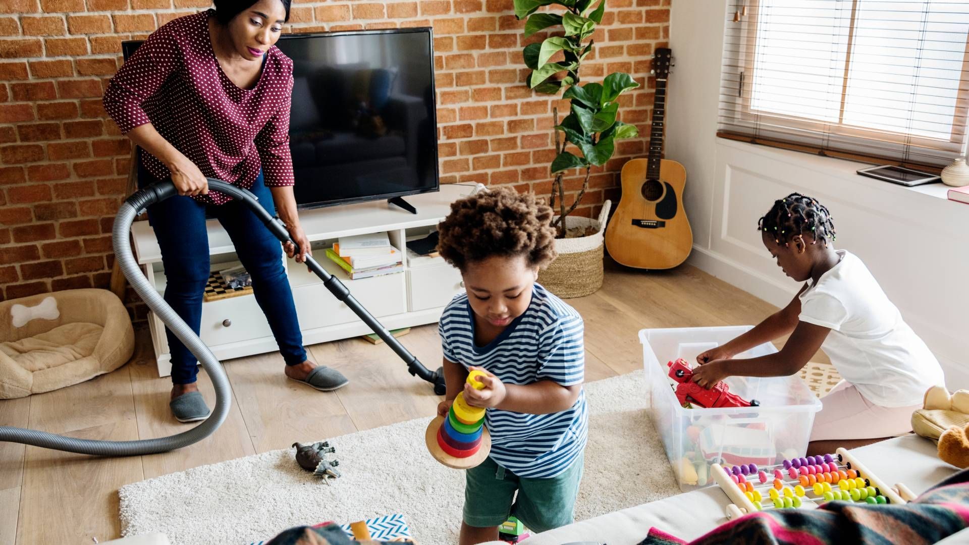 A woman is vacuuming the floor while two children play on the floor.