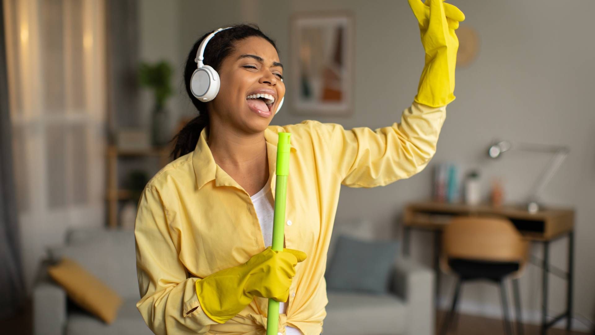 A woman wearing headphones and holding a mop is dancing in a living room.