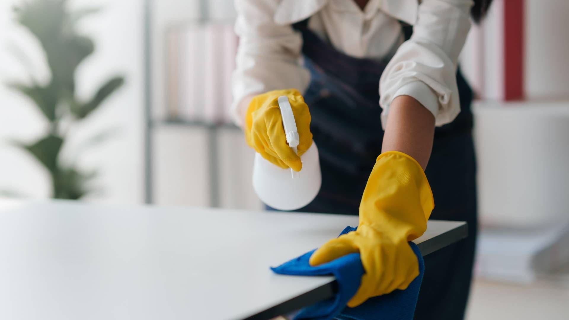 A woman wearing yellow gloves is cleaning a table with a cloth and spray bottle.