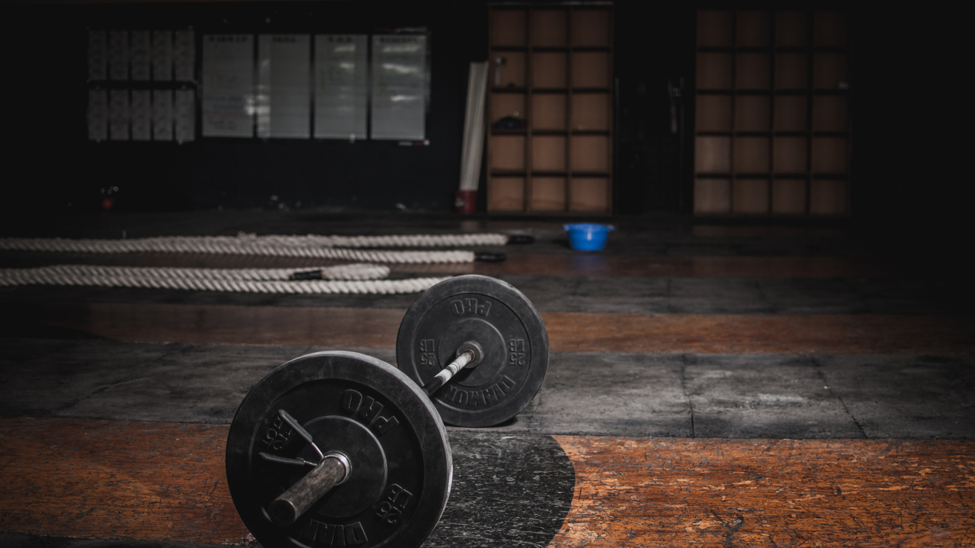 Barbells on a gym floor, with ropes and blue bucket. Dark interior, focused lighting.