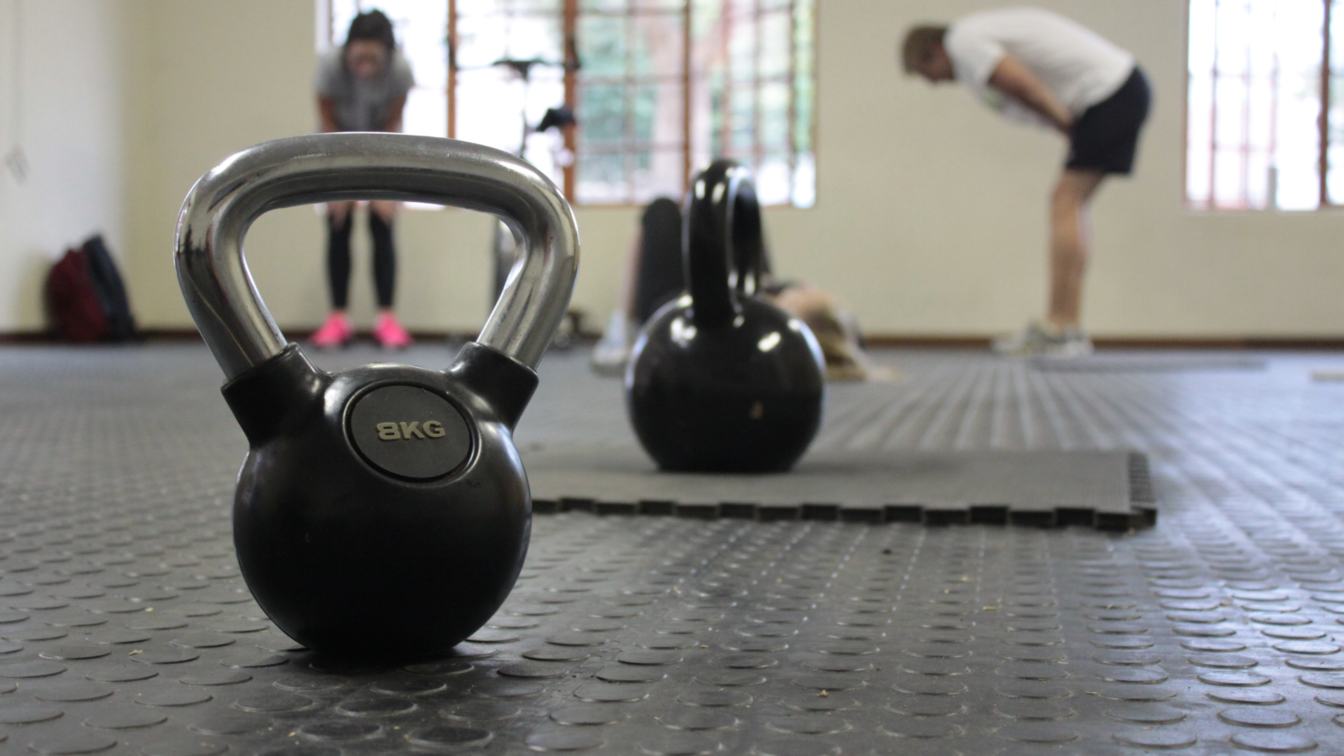 Two kettlebells on the floor of a gym, people exercising in the background.