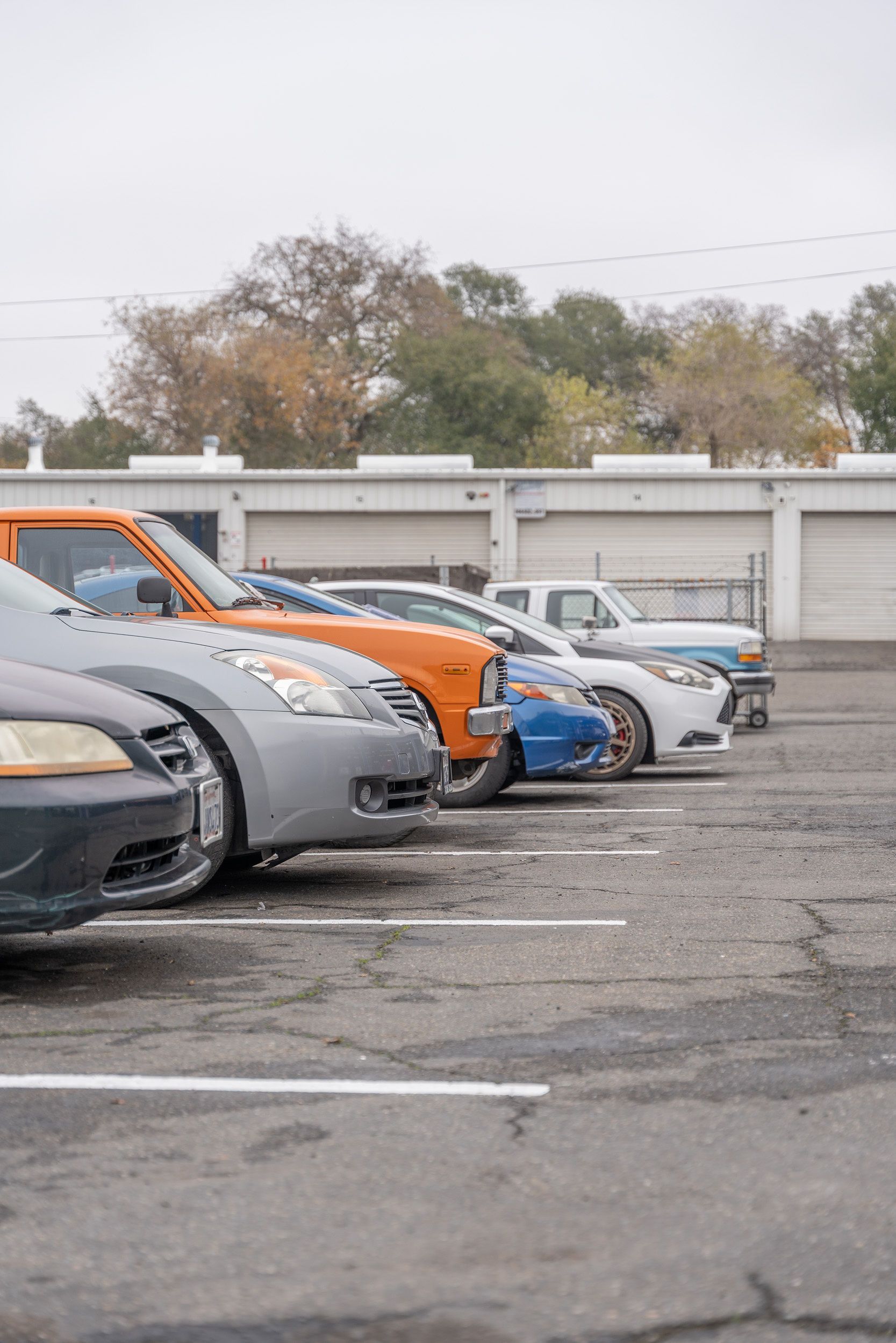 Cars parked in a line in a parking lot; a few are different colors, including orange and blue.