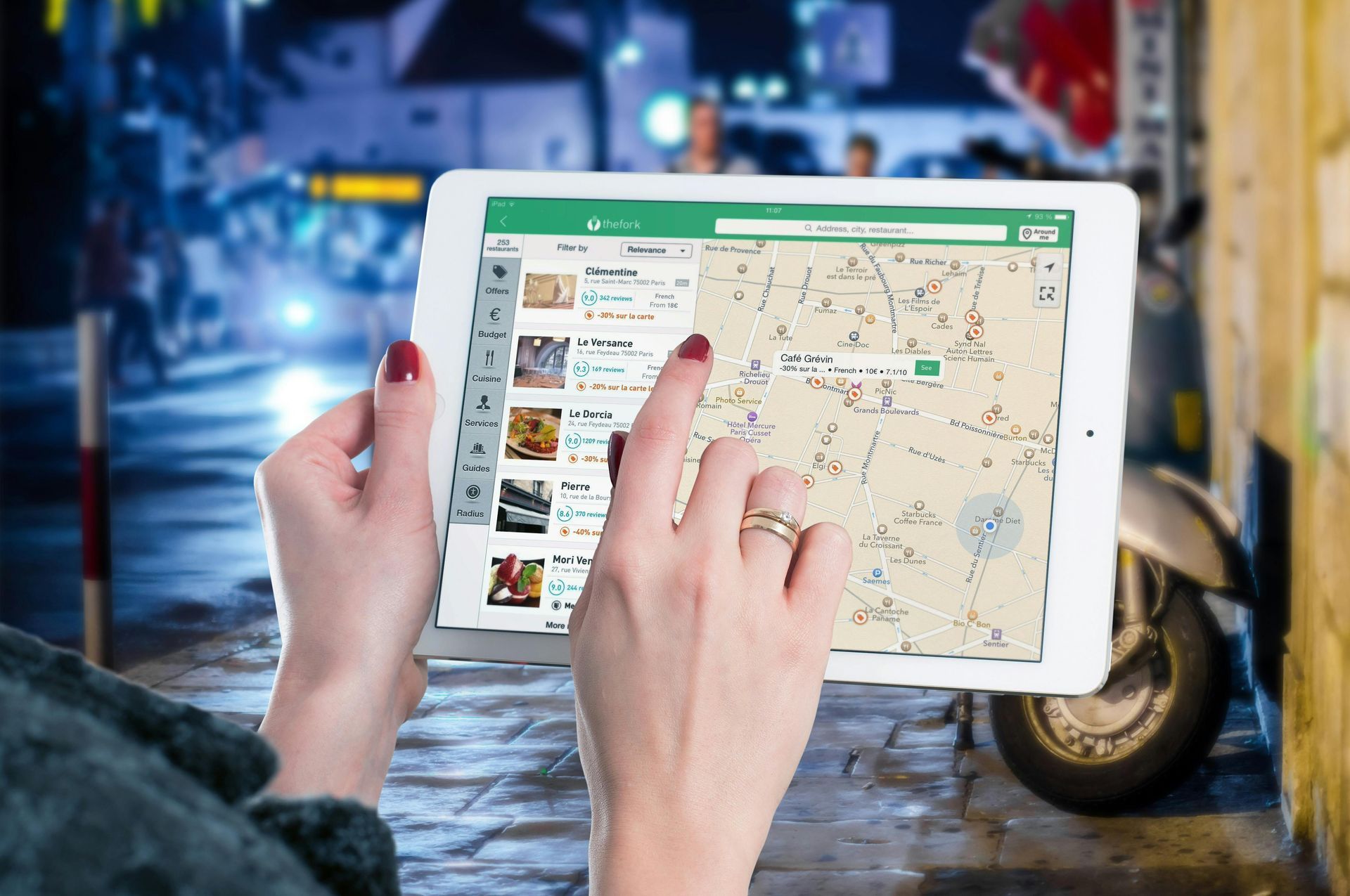 Woman uses a tablet with a map app outdoors at night, street and vehicle in the background.