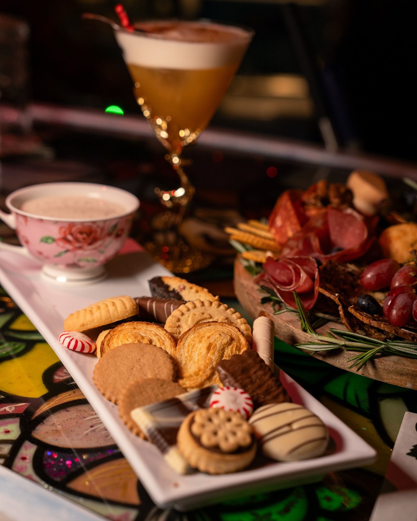 Cocktail, cookies, teacup, and charcuterie board on a table. Dark setting with colorful accents.