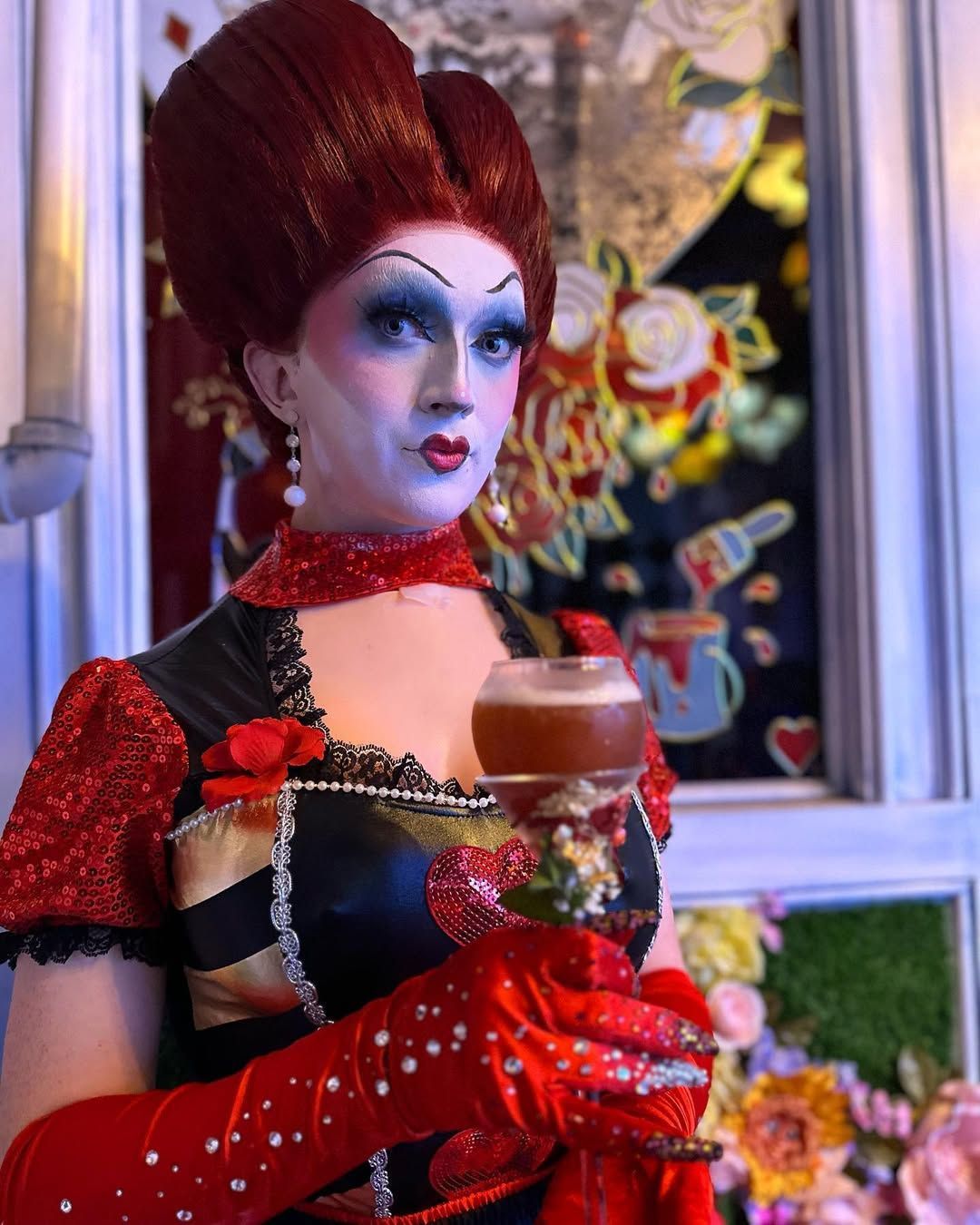A drag queen dressed as the Queen of Hearts, holding a drink, in front of a floral backdrop.