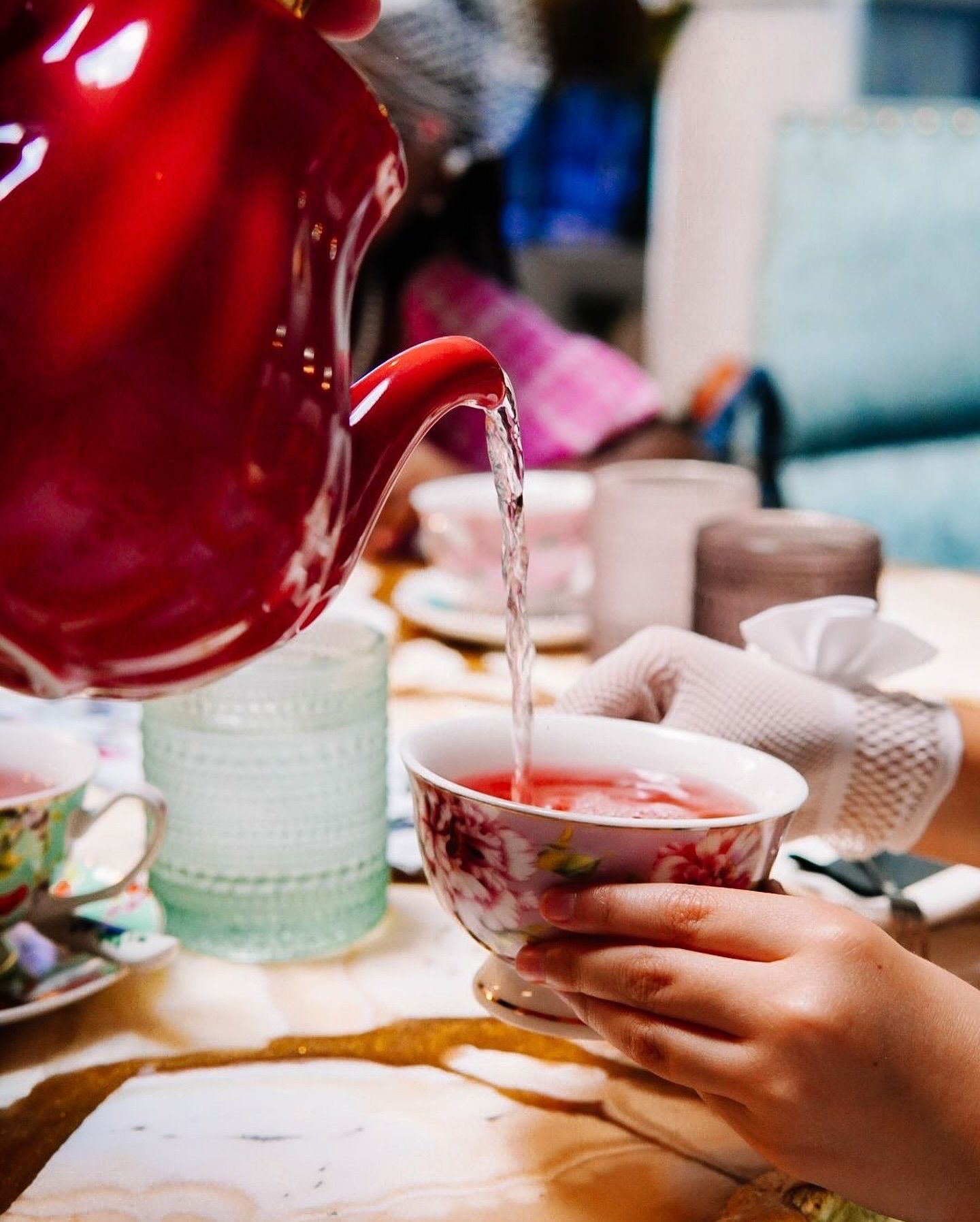 Red teapot pouring tea into floral teacup held by gloved hands at a table.