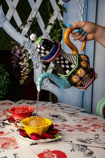 Hand pouring from a decorated teapot into a yellow teacup. The table is set for tea with a red and white tablecloth.