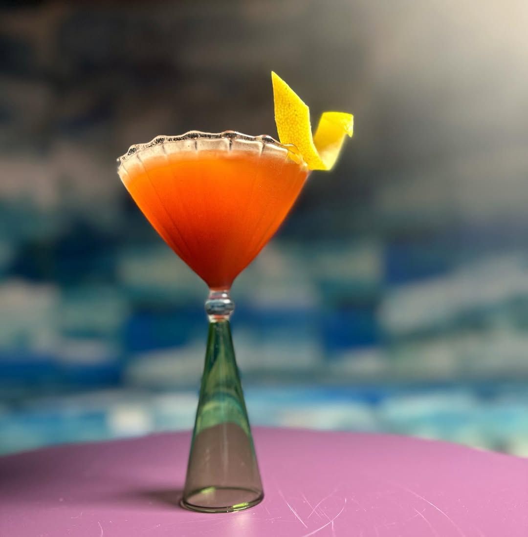 Orange cocktail in a stemmed glass, lemon peel garnish, salt rim, set on pink table, blurred background.
