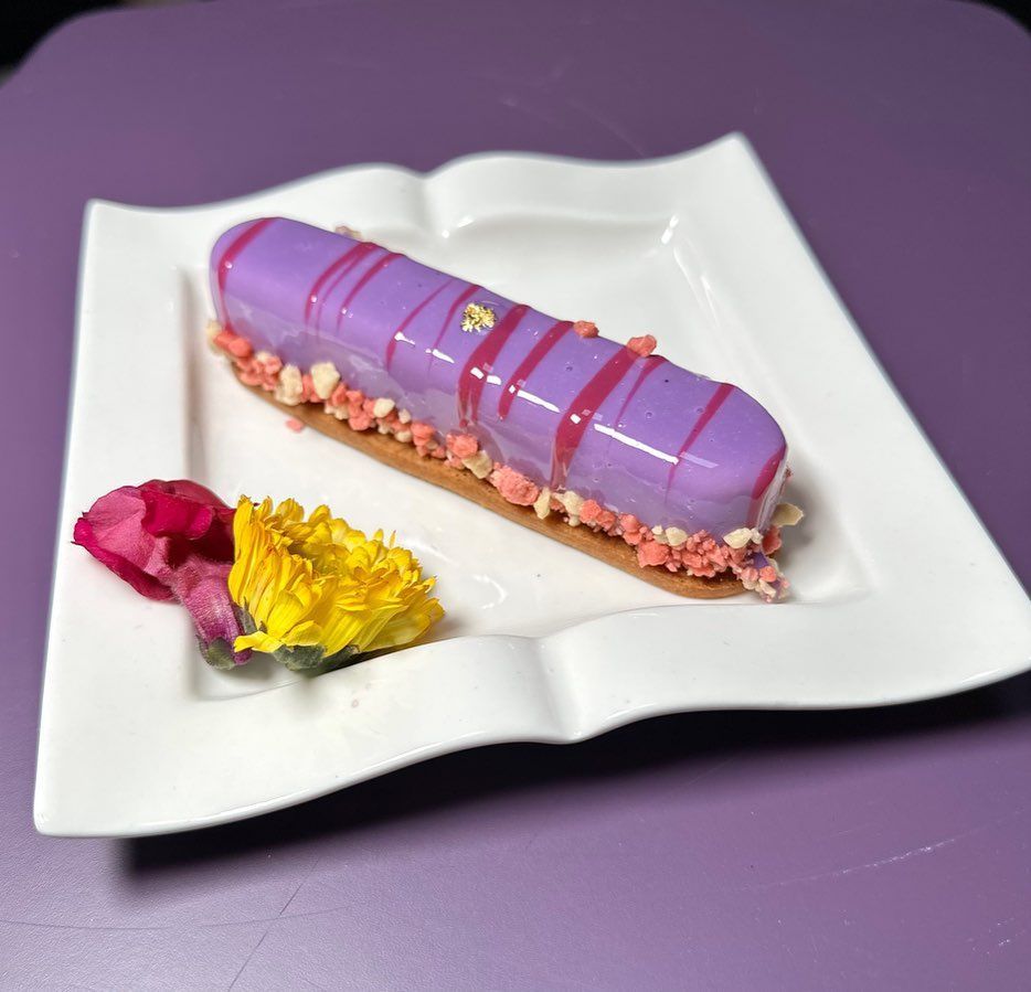 A purple glazed eclair on a white plate, decorated with pink accents, yellow and red flowers.