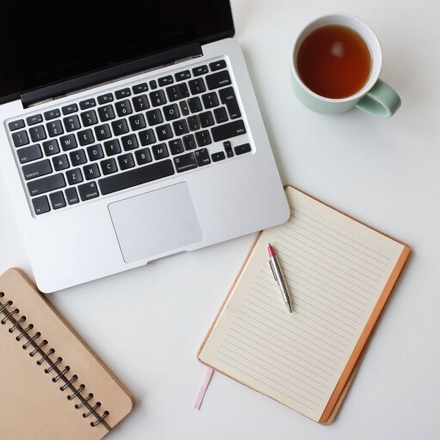 Laptop, notitieboekjes, pen en een mok thee, uitgestald op een witte ondergrond.