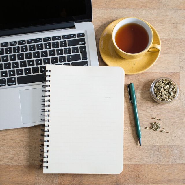 Laptop, notitieblok, pen, theekopje en een klein schaaltje met kruiden op een houten bureau.