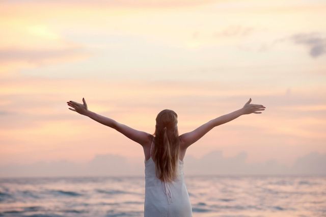 Vrouw met uitgestrekte armen, kijkend naar de zonsondergang boven de oceaan.