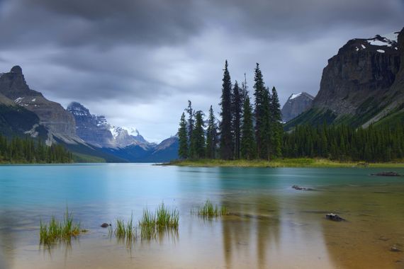 Een turquoise meer met bergen, donkere wolken en een klein eilandje met bomen.