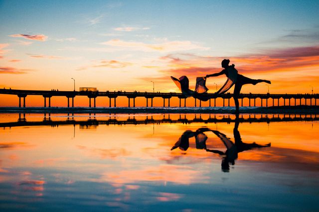 Silhouet van een danseres die een doek vasthoudt, in evenwicht in ondiep water, weerspiegeld tegen de zonsondergang; pier.