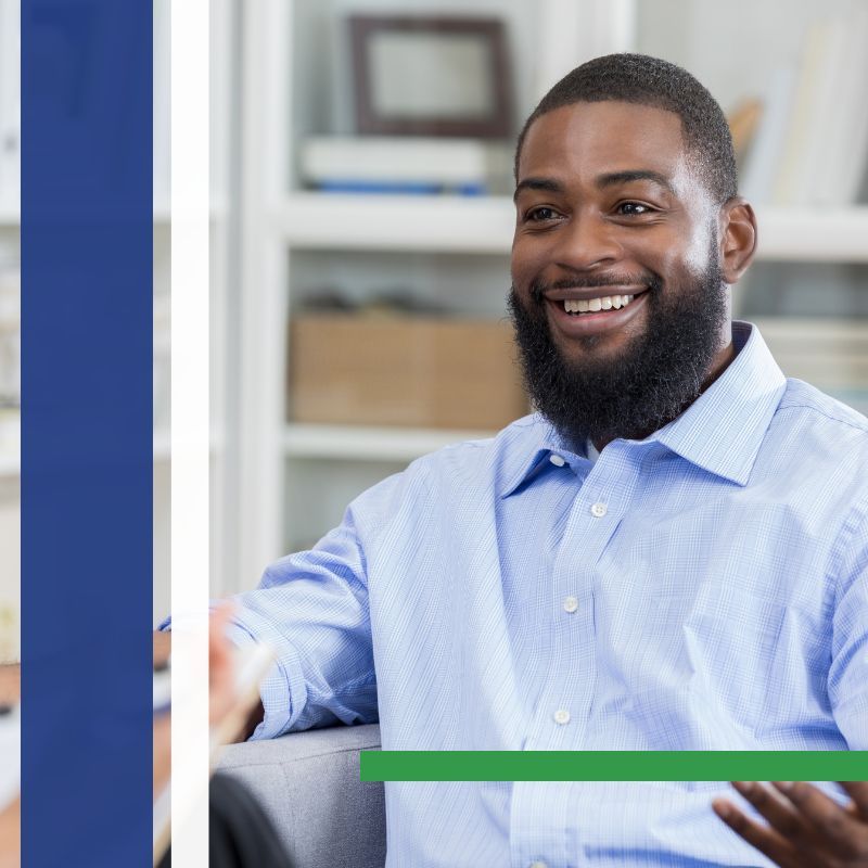 Smiling man with a beard gestures while seated, wearing a light blue button-down shirt.