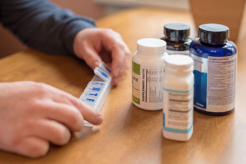 Person opening a pill organizer, several supplement bottles on a wooden table.