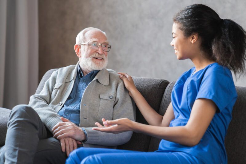 Woman in blue scrubs comforts elderly person on a couch. The man wears glasses and a jacket.