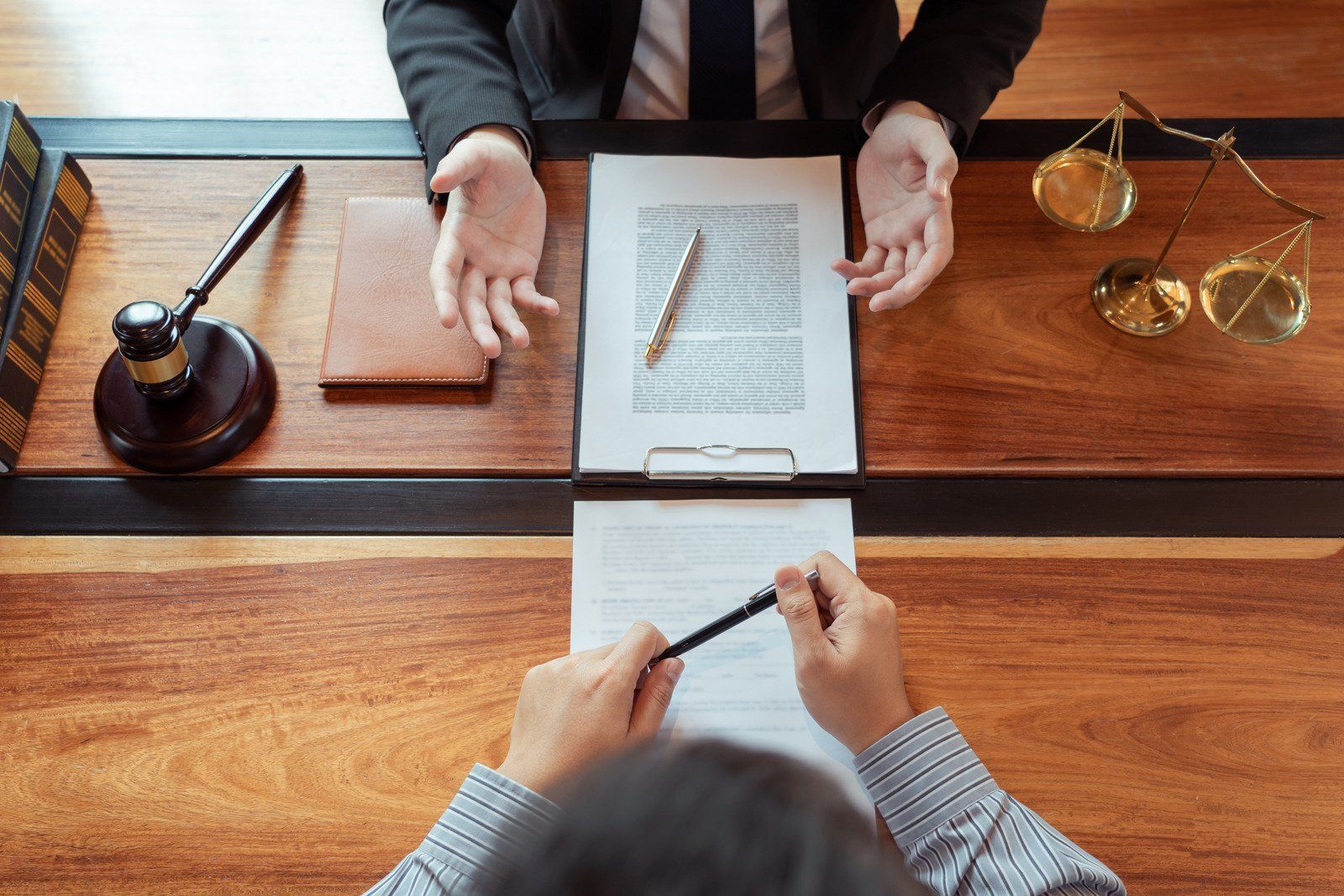 Lawyer gestures while discussing document with client at a wooden table. Gavel and scales of justice visible.