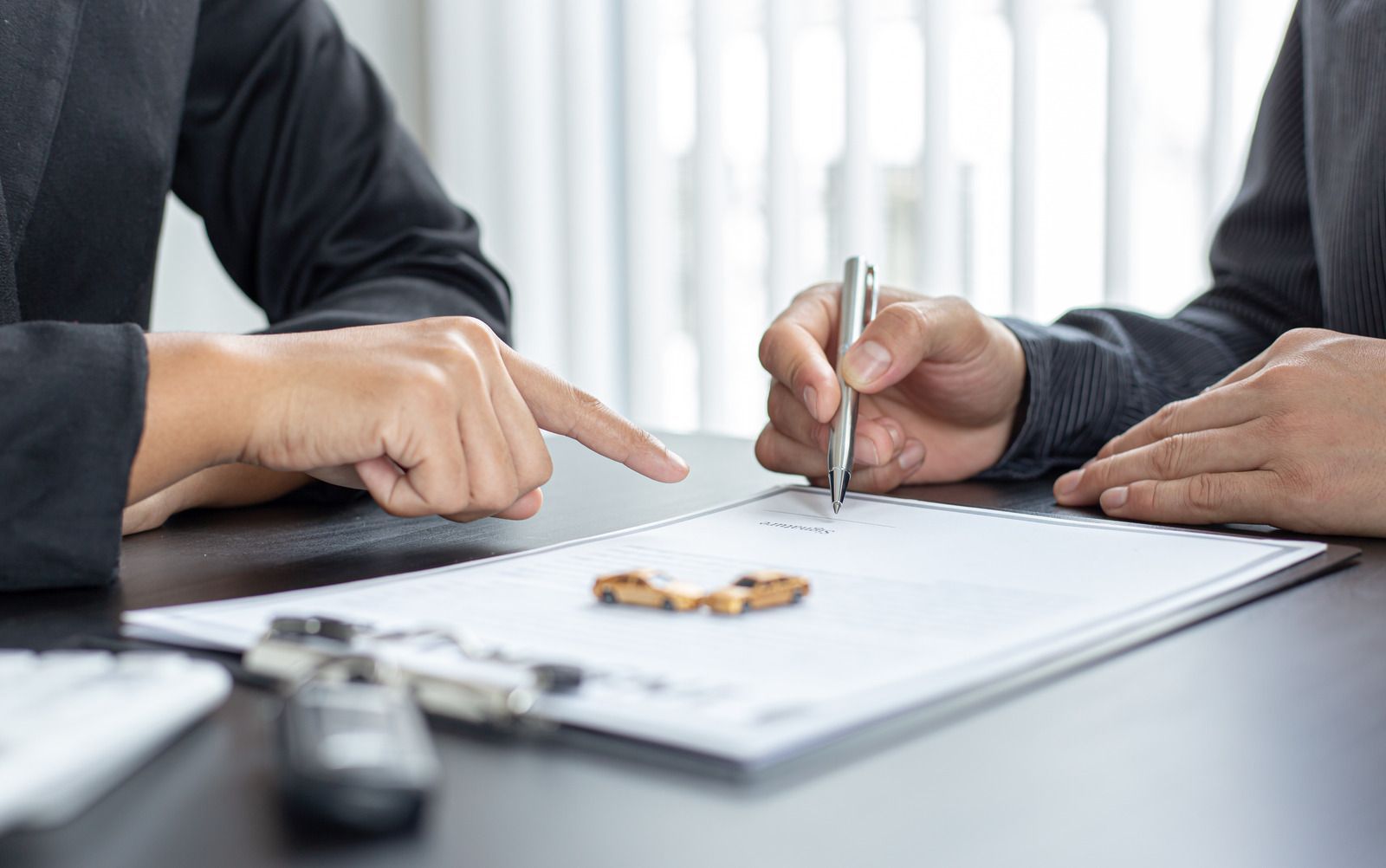 Two people in suits at a desk, one signing a document, car keys and a car on the paper.