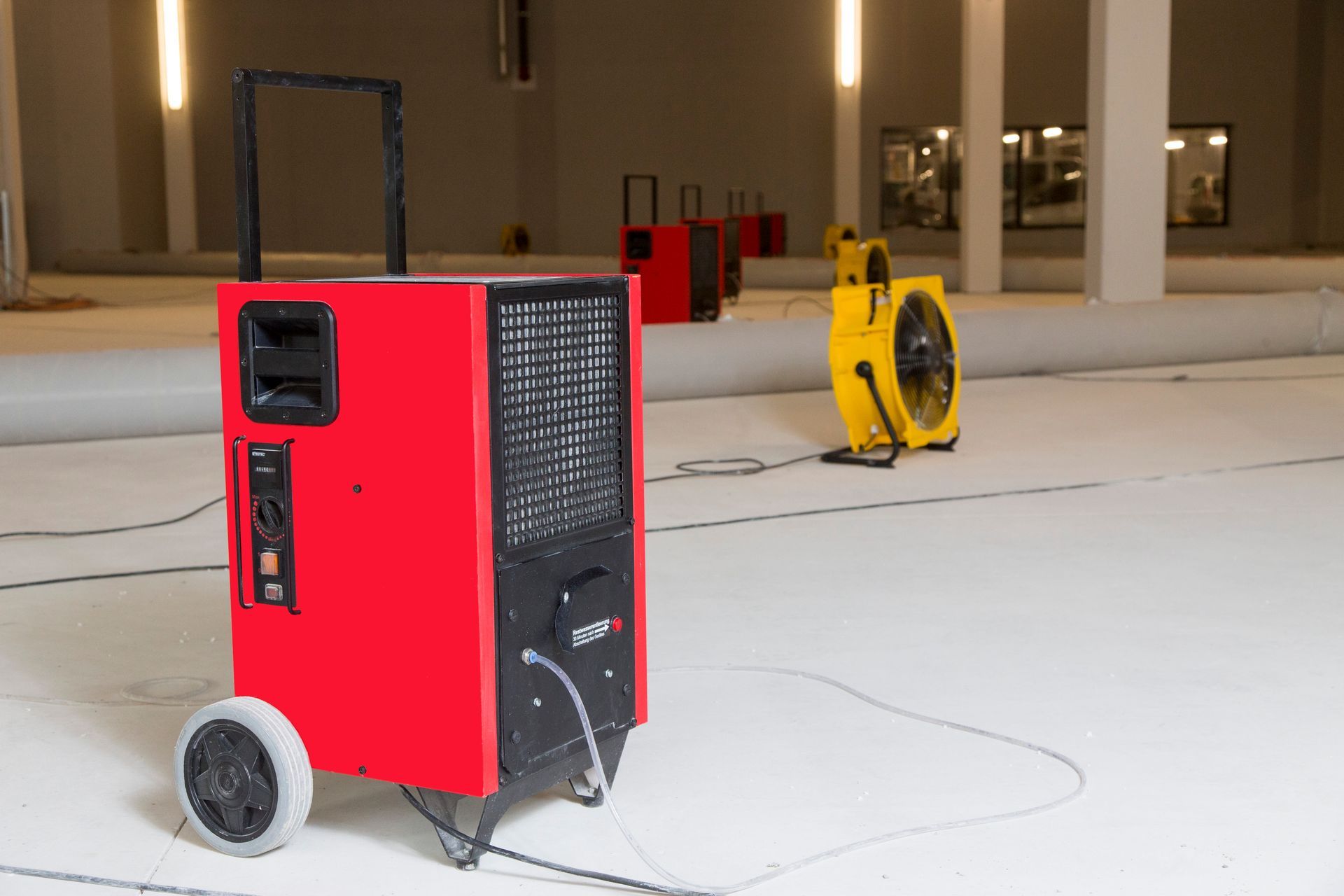 Roter Luftentfeuchter auf Rädern in einem weissen Raum, mit einem gelben Ventilator und anderen Gerä