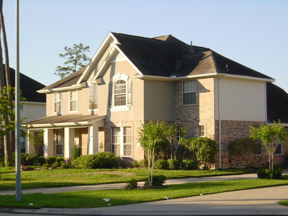 Two-story house with beige and brick siding, green lawn, and shrubs in front.