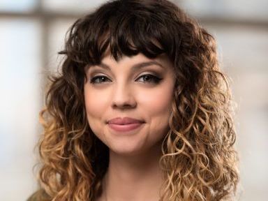 A woman with curly hair is wearing a green sweater and a necklace.