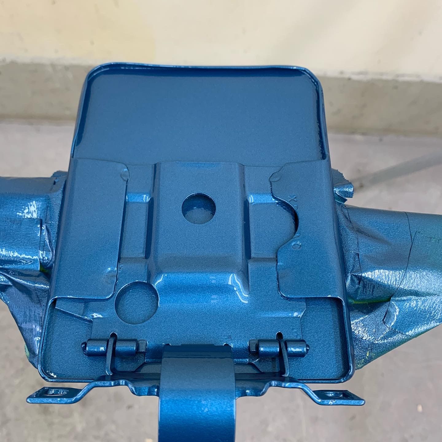 Blue metal hinged bracket mounted on a pipe, shown close up against a light wall.