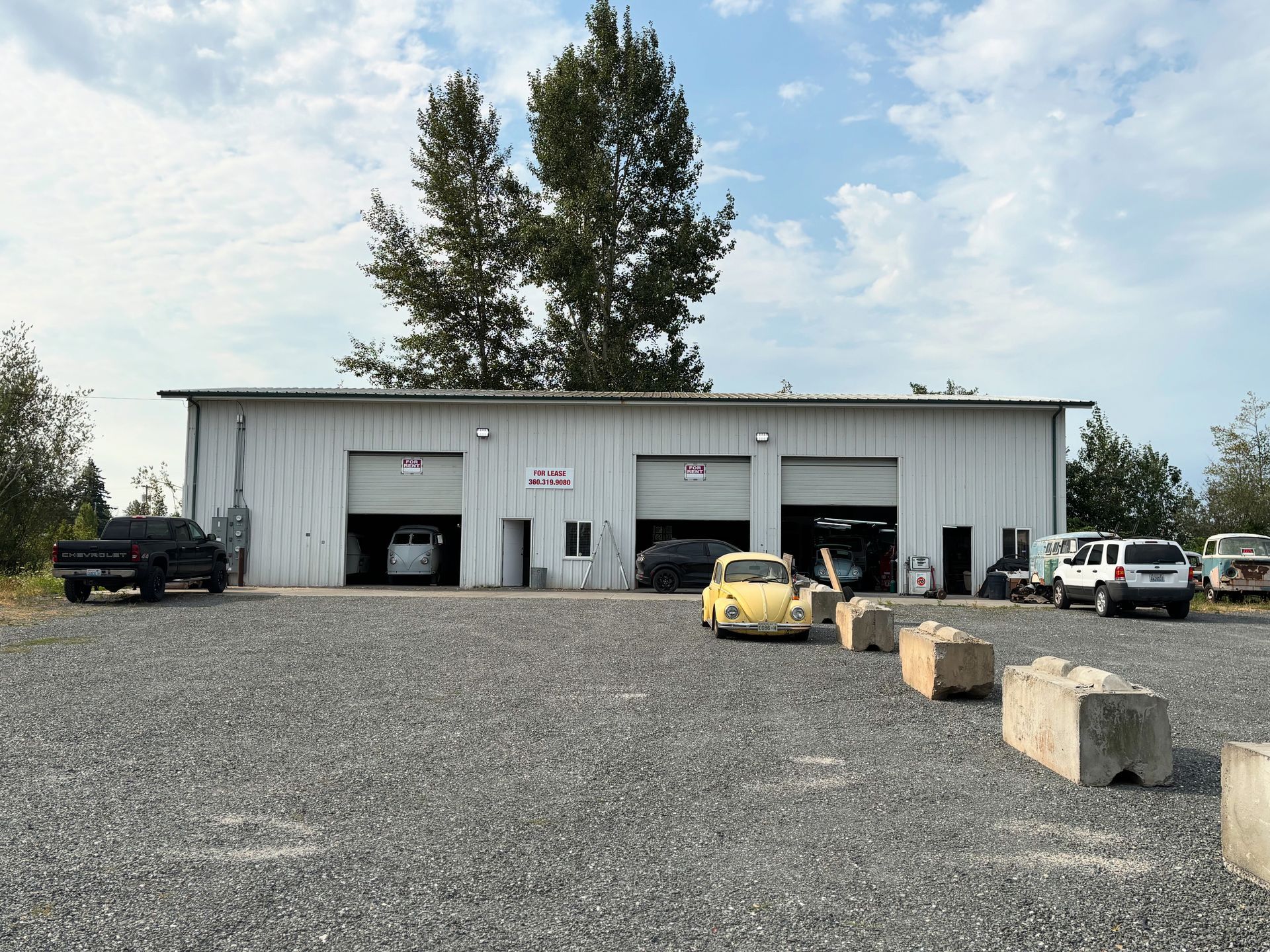 Gravel lot outside a white garage building with trucks, cars, and concrete blocks in front